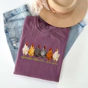 May include: A plum-colored t-shirt featuring a graphic of six chickens in various colors standing on a green patch. The shirt is layered over light blue denim jeans and accompanied by a tan wide-brimmed hat and a delicate gold chain necklace.