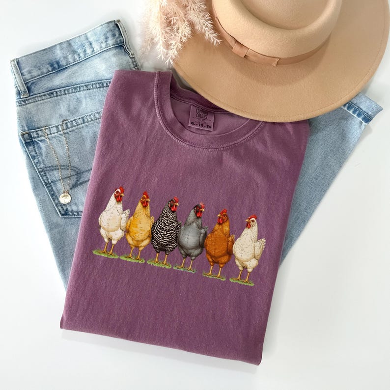 May include: A plum-colored t-shirt featuring a graphic of six chickens in various colors standing on a green patch. The shirt is layered over light blue denim jeans and accompanied by a tan wide-brimmed hat and a delicate gold chain necklace.
