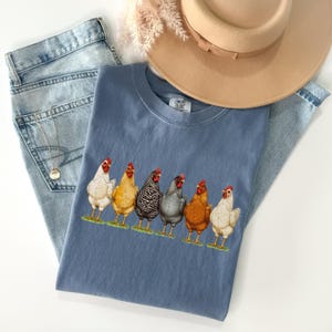 May include: A blue t-shirt featuring a graphic of six chickens in various colors including white, yellow, black and white striped, grey, and brown, standing on a green patch. The shirt is paired with light blue denim jeans and a tan hat.