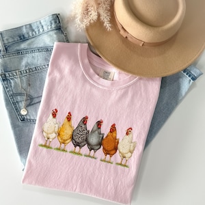 May include: A light pink t-shirt featuring a graphic of six chickens in various colors standing in a row on a green patch of grass. The shirt is layered with light blue denim jeans, a tan wide-brimmed hat, and a thin silver chain necklace.