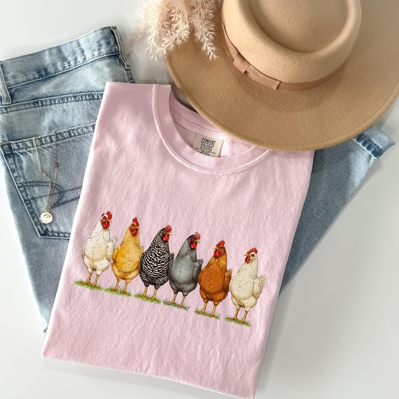 May include: A light pink t-shirt featuring a graphic of six chickens in various colors standing in a row on a green patch of grass. The shirt is layered with light blue denim jeans, a tan wide-brimmed hat, and a thin silver chain necklace.
