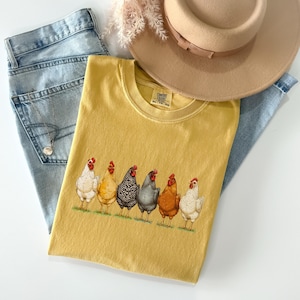 May include: A mustard yellow t-shirt featuring a graphic print of six chickens in various colors standing in a row. The shirt has a crew neckline and a Comfort Colors label. It is displayed with light blue denim jeans and a tan wide-brimmed hat.