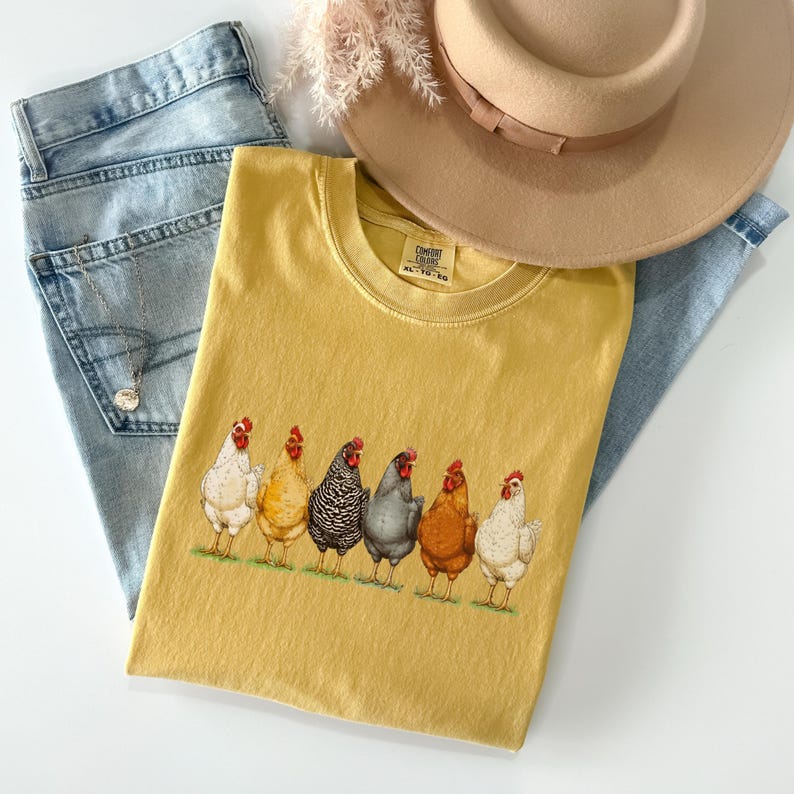 May include: A mustard yellow t-shirt featuring a graphic print of six chickens in various colors standing in a row. The shirt has a crew neckline and a Comfort Colors label. It is displayed with light blue denim jeans and a tan wide-brimmed hat.