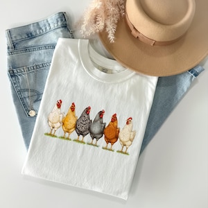May include: A white t-shirt featuring a graphic print of six chickens in various colors including white, yellow, black and white striped, grey, and brown. The shirt is layered with light blue denim jeans, a tan wide-brimmed hat, and a silver necklace.