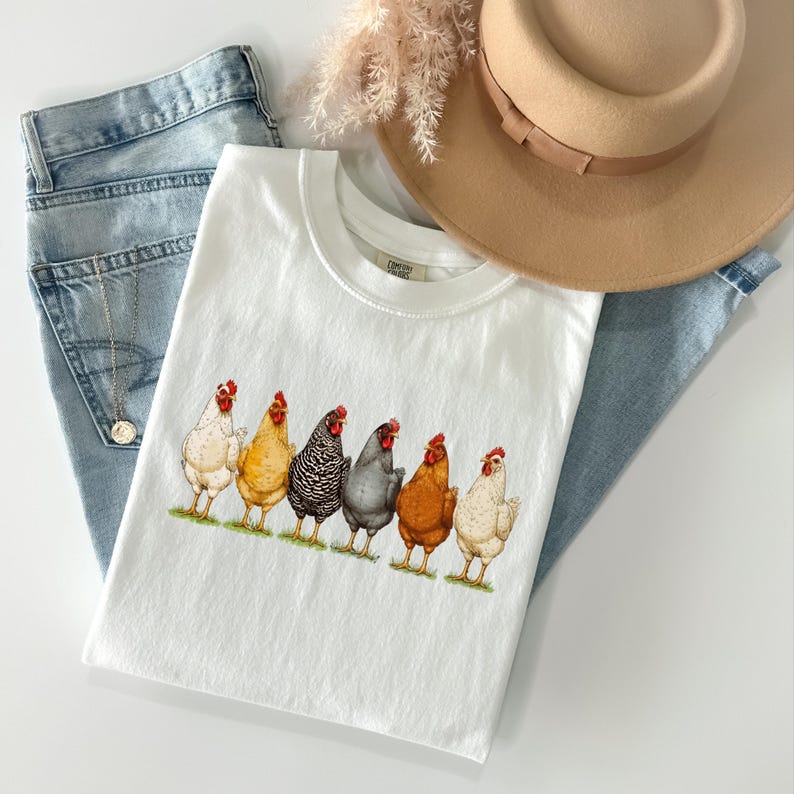 May include: A white t-shirt featuring a graphic print of six chickens in various colors including white, yellow, black and white striped, grey, and brown. The shirt is layered with light blue denim jeans, a tan wide-brimmed hat, and a silver necklace.