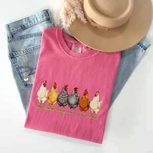 May include: A bright pink short-sleeved t-shirt featuring a graphic print of six chickens in various colors including white, yellow, black and white speckled, grey, and brown, standing in a row on a thin strip of green grass.