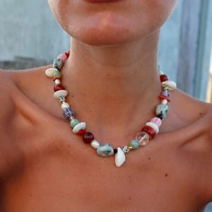 Puede incluir: Un collar de cuentas de colores con una variedad de piedras y formas. El collar incluye cuentas rojas, blancas, verdes y rosas, con un colgante de concha blanca. El collar se lleva alrededor del cuello.