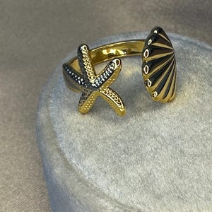 Peut inclure: Bague dorée ornée d'un motif d'étoile de mer et de coquillage. L'étoile de mer présente une surface texturée avec des accents noirs, tandis que le coquillage arbore des rayures noires. La bague ouverte est réglable et est présentée sur une surface grise douce.