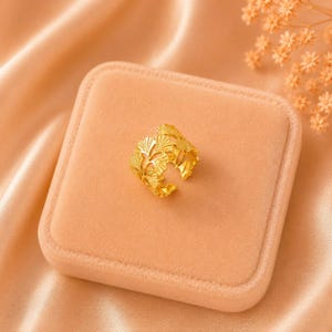 May include: A gold-colored ring with a leaf design, resting on a peach-colored velvet surface. The ring's intricate details are visible, showcasing a nature-inspired aesthetic. The ring is a piece of jewelry.