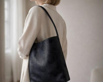 Puede incluir: Un bolso tote de cuero negro con una correa larga para el hombro. El bolso tiene una superficie texturizada y un diseño simple y minimalista. El bolso se lleva al hombro.