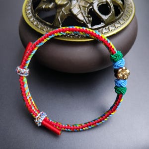 May include: A colorful braided bracelet with red, yellow, blue, and green threads. The bracelet features a gold-colored flower charm and two silver-colored rhinestone accents. The bracelet is a traditional style.