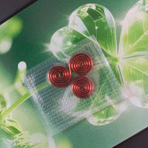 Puede incluir: Un pequeño componente de alambre metálico rojo formado por tres espirales interconectadas que crean un patrón similar a un trisquel. El objeto está encerrado en una bolsa de plástico transparente y texturizada, resaltando el acabado rojo vibrante.