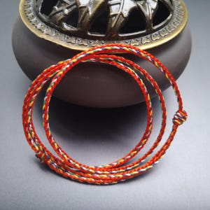 Può includere: Un cordoncino intrecciato rosso con accenti colorati, annodato a un'estremità. Il cordoncino è avvolto in un motivo circolare, con un vaso di ceramica marrone e un oggetto metallico decorativo sullo sfondo.