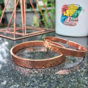 May include: Two copper cuff bracelets with a hammered, textured finish. The bracelets are circular with an open gap design, showcasing a metallic reddish-brown color and a reflective surface that catches light across the uneven, dimpled metal texture.