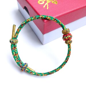 Può includere: Un braccialetto intrecciato con fili verdi, gialli, rossi e dorati. Il braccialetto presenta nodi decorativi e dettagli metallici color oro. Il braccialetto è esposto vicino a una scatola regalo rossa con scritte dorate.