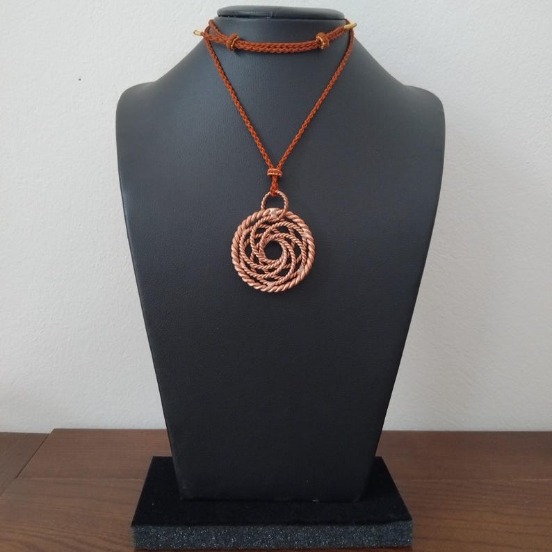 May include: A copper-colored circular pendant featuring a complex, interwoven rope-like design. The pendant is suspended from a thin, braided rust-colored cord that wraps around the neck, secured with small metallic beads at the back of the cord.