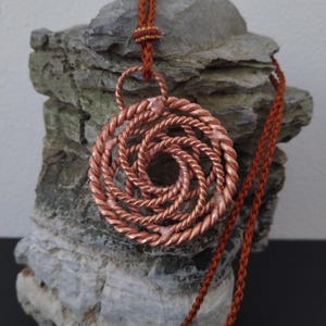 May include: A circular pendant crafted from twisted copper wire, featuring a spiral pattern that radiates from the center. The pendant is suspended from a rust-colored braided cord, which includes a small decorative knot detail above the pendant.