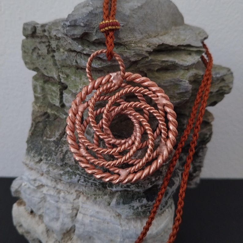 May include: A circular pendant crafted from twisted copper wire, featuring a spiral pattern that radiates from the center. The pendant is suspended from a rust-colored braided cord, which includes a small decorative knot detail above the pendant.