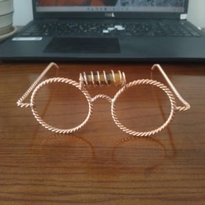 May include: A pair of round glasses crafted from twisted copper wire, featuring a bridge adorned with a horizontal, cylindrical tiger's eye gemstone wrapped in a copper wire coil. The frame has thin, twisted wire temples extending outward.