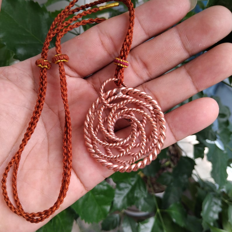 May include: A circular pendant crafted from twisted copper wire, featuring a complex spiral pattern. The pendant is suspended from a braided, rust-colored cord that includes adjustable sliding knots and gold-toned metal tips at the ends.