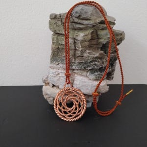 May include: A circular pendant crafted from twisted copper wire, featuring an intricate spiral pattern. The pendant hangs from a thin, braided rust-colored cord that includes adjustable sliding knots and a small metallic tip at the end of the cord.