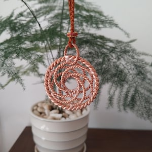 May include: A circular pendant crafted from twisted copper wire, featuring a spiraling, open-work design. The pendant is suspended from a braided brown cord that includes a small decorative knot with red and gold thread accents at the top.