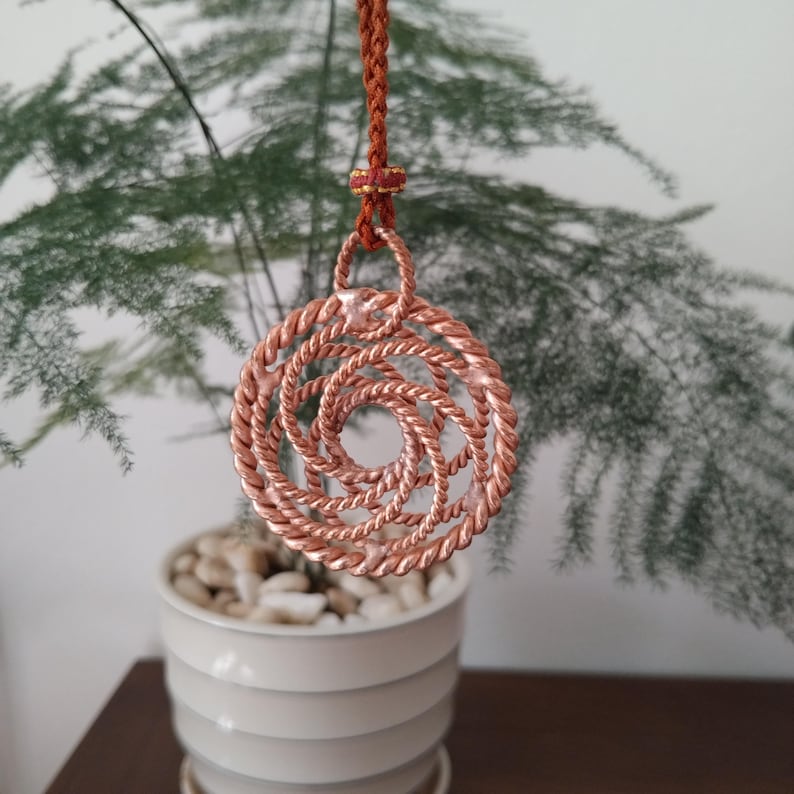 May include: A circular pendant crafted from twisted copper wire, featuring a spiraling, open-work design. The pendant is suspended from a braided brown cord that includes a small decorative knot with red and gold thread accents at the top.