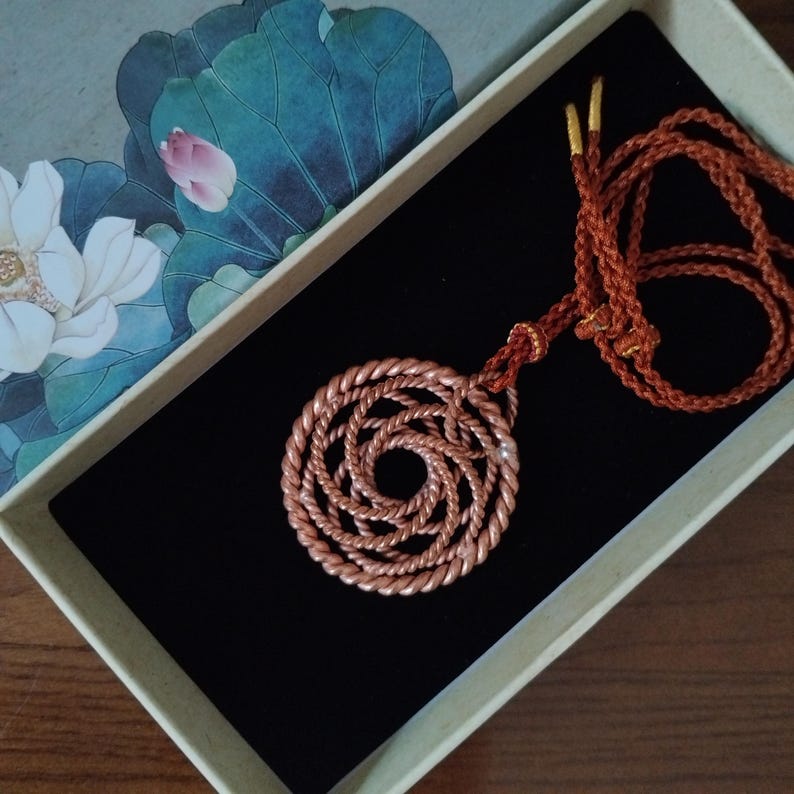 May include: A copper-colored pendant featuring a circular, spiraling rope-like design attached to a matching braided cord. The pendant is presented inside a rectangular box with a lid decorated with a white lotus flower and green leaves.