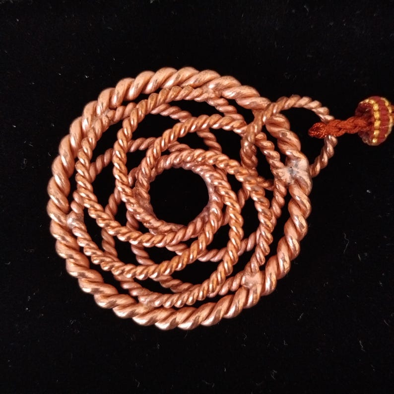 May include: A circular pendant crafted from twisted copper wire arranged in a spiral pattern. The metallic copper surface has a warm, reddish-brown tone. A small, textured, reddish-brown fabric loop is attached to the top of the pendant.