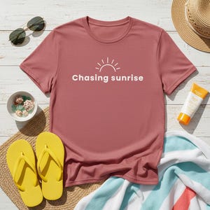 May include: A dusky pink t-shirt with the words "Chasing sunrise" and a sun graphic. The image includes yellow flip-flops, sunglasses, a straw hat, sunscreen, and a small bowl with shells, suggesting a beach theme.