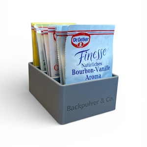 May include: A grey rectangular container holding several packets of baking ingredients. The packets are various colours, including yellow, white, and blue. The front packet reads "Dr. Oetker Finesse Natürliches Bourbon-Vanille Aroma". The container has the text "Backpulver & Co." on the side.