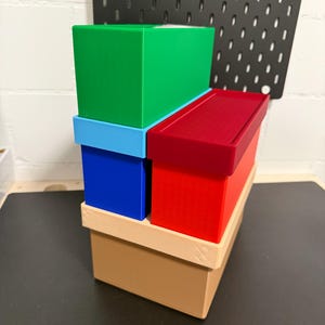 Könnte beinhalten: Ein Stapel bunter rechteckiger Boxen. Die Boxen sind in den Farbtönen Grün, Blau, Rot und Beige gehalten. Die Boxen sind aufeinander gestapelt, mit der größten beigefarbenen Box unten und der grünen Box oben. Die Boxen bestehen aus einem festen Material.