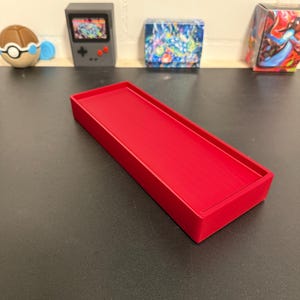 Könnte beinhalten: Ein rechteckiges, leuchtend rotes Tablett mit erhöhten Kanten. Das Tablett besteht aus einem festen Material und steht auf einer dunklen Oberfläche. Im Hintergrund befinden sich kleine dekorative Gegenstände, darunter eine Miniatur-Spielkonsole und eine Pokémon-Figur.