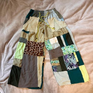 May include: Patchwork trousers with a drawstring waist. The trousers feature a variety of fabrics in shades of green, brown, tan, and leopard print. The wide-leg design is made from various fabric patches.