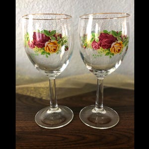 May include: Two clear wine glasses with a floral rose design in red, yellow, and pink. The rims of the glasses are trimmed with gold. The glasses are on a dark wooden surface.