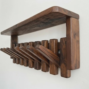 Puede incluir: Un perchero de pared de madera marrón oscuro con una estantería. El perchero tiene múltiples clavijas anguladas para colgar objetos. La veta de la madera es visible y el diseño es minimalista y funcional.