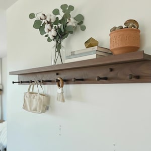 May include: A wooden shelf with a row of pegs, holding a small canvas tote bag and a wooden ring with a tassel. The shelf also displays a glass vase with cotton stems, books, and a terracotta pot with mushrooms. The shelf is mounted on a white wall.