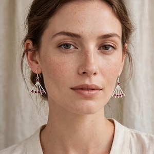 May include: A pair of silver dangle earrings featuring a fan-shaped design with five radiating metal spokes, each tipped with a small, round, deep pink gemstone. The earrings hang from simple silver hooks and have a polished metallic finish.