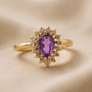 May include: A gold ring featuring a large oval purple gemstone surrounded by smaller clear stones. The ring's band is a simple, polished gold. The setting of the ring is a starburst design, with the purple stone as the focal point.
