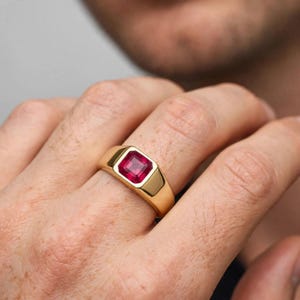 May include: A gold signet ring featuring a square-cut, deep red gemstone set in the center. The band is smooth and polished, with a wide, flat top surface that frames the vibrant red stone, creating a bold and classic jewelry piece.