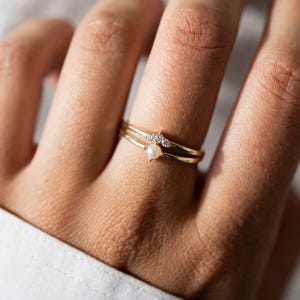 May include: A thin gold ring featuring a small, round white pearl centered on the band, with a delicate V-shaped accent above the pearl set with three tiny, clear, sparkling stones. The metal band is smooth, polished, and yellow in color.