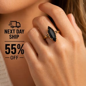 Op de afbeelding: Een gouden ring met een zwarte edelsteen in marquise-slijpvorm in een klauwzetting. De ringband heeft een gespleten ontwerp waar deze de centrale steen ontmoet. De ring wordt om een vinger gedragen en toont de langwerpige vorm van de steen.