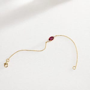 Könnte beinhalten: Ein zartes Armband aus einer goldfarbenen Kette mit einem zentralen, marquise-geschliffenen dunkelroten Edelstein, der von zwei kleinen klaren Akzentsteinen flankiert wird. Das Armband besitzt eine feine Gliederkette und einen Karabinerverschluss.