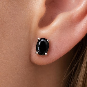 May include: An oval-shaped black gemstone stud earring set in a four-prong silver-toned metal setting. The faceted stone is positioned centrally on the earlobe, showcasing a polished surface and a deep, dark color with reflective highlights.