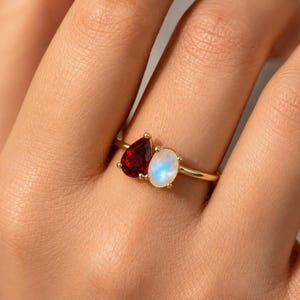 May include: A gold ring featuring a pear-shaped red gemstone and an oval-shaped, iridescent white gemstone. The ring is set on a thin gold band.