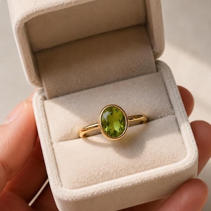 May include: A gold ring featuring an oval-cut, light green peridot gemstone set in a smooth bezel setting. The ring is presented inside a beige, velvet-lined jewellery box with a hinged lid, showcasing the polished metal band and faceted stone.