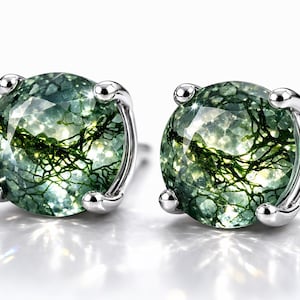 Pode incluir: Um par de brincos redondos com pedras de ágata musgo facetadas com intrincadas inclusões dendríticas verde-escuras, montadas em garras de metal prateado. As pedras têm uma qualidade translúcida com uma superfície facetada cintilante.