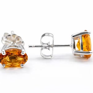 Könnte beinhalten: Ein Paar Ohrstecker mit ovalen, goldgelben Edelsteinen, die in einem silberfarbenen Metallrahmen gefasst sind. Die Ohrringe werden vor einem weißen Hintergrund präsentiert, um ihr Design und ihre Farbe hervorzuheben.