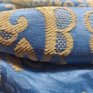 May include: Close-up of folded blue fabric with a gold floral pattern. The fabric has a soft, velvety texture and a subtle sheen. The gold pattern is raised, creating a textured effect. The overall design is elegant and luxurious.