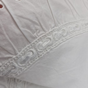 May include: Close-up of white fabric with eyelet details and a decorative embroidered border. The fabric appears to be cotton or a similar lightweight material, with a slightly textured surface. The design suggests a garment or textile.